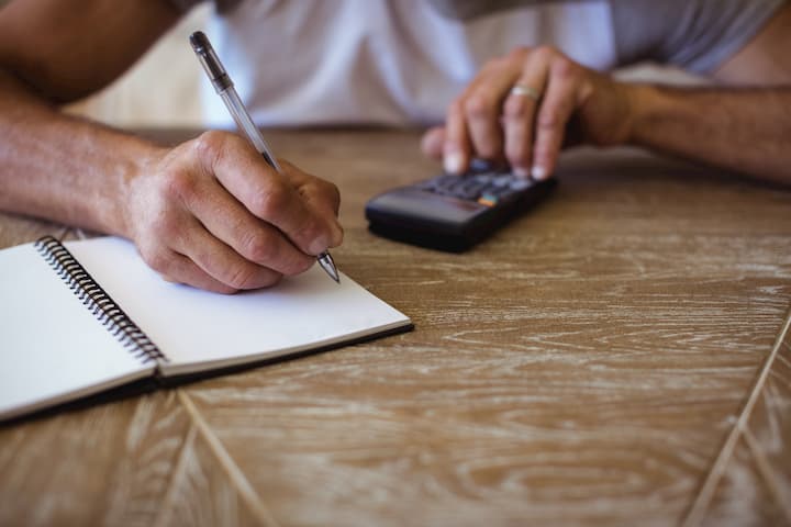 homem usando uma calculadora em uma mesa de madeira. Ao mesmo tempo em que faz as contas, anota os resultados em um caderno